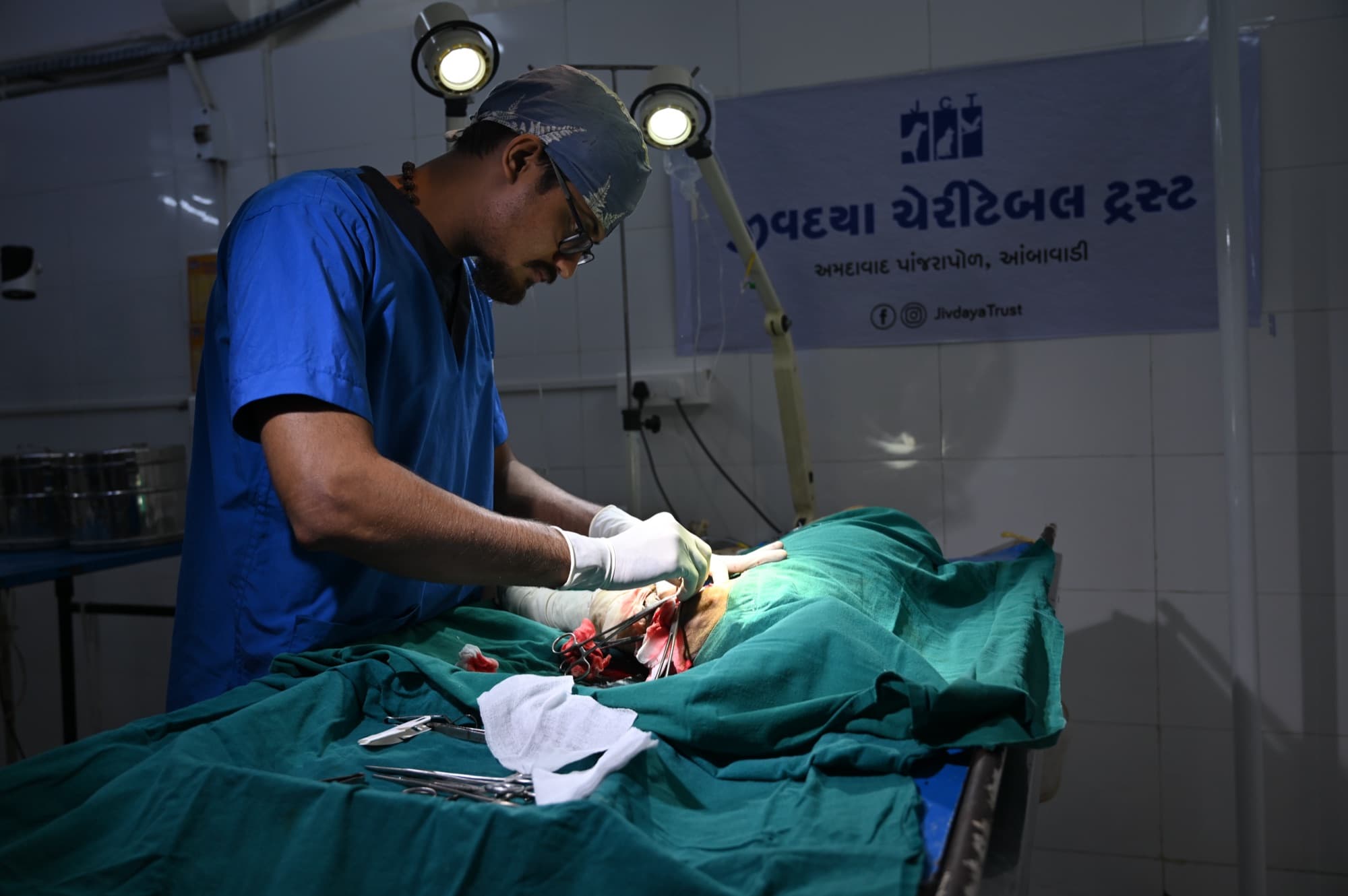 A surgeon wearing a headlamp operating on a draped patient beneath a Jivdaya Charitable Trust banner